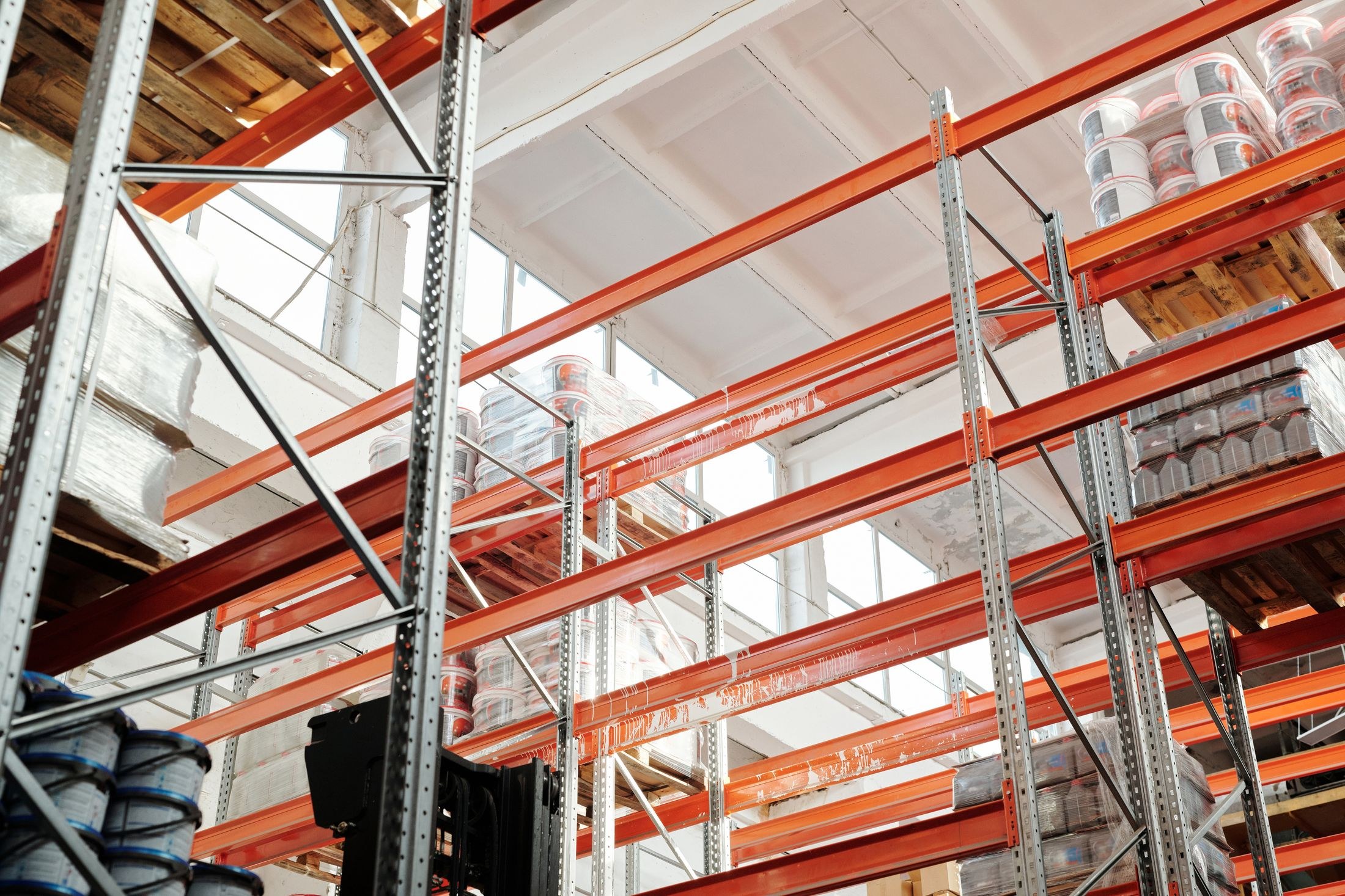 Tall orange and steel warehouse racks hold wooden pallets of wrapped containers in a bright industrial interior